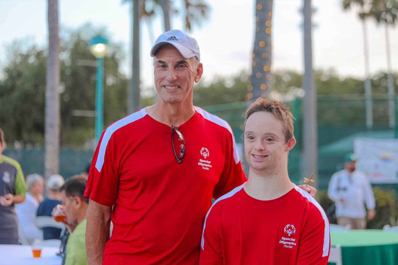 A coach and a beaming Special Olympics athlete standing together under the palms