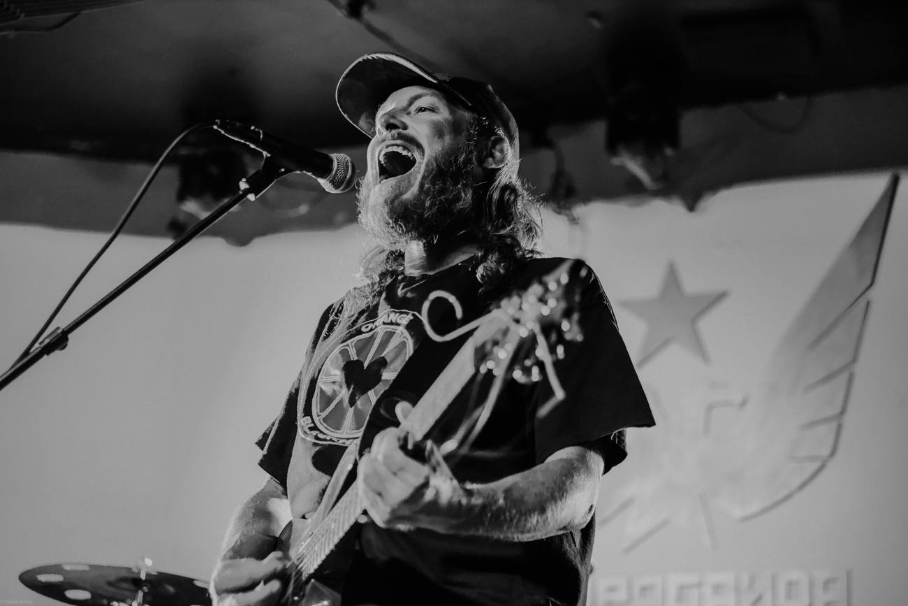 Goat Rope's frontman mid-howl at the microphone, guitar in hand, in black and white