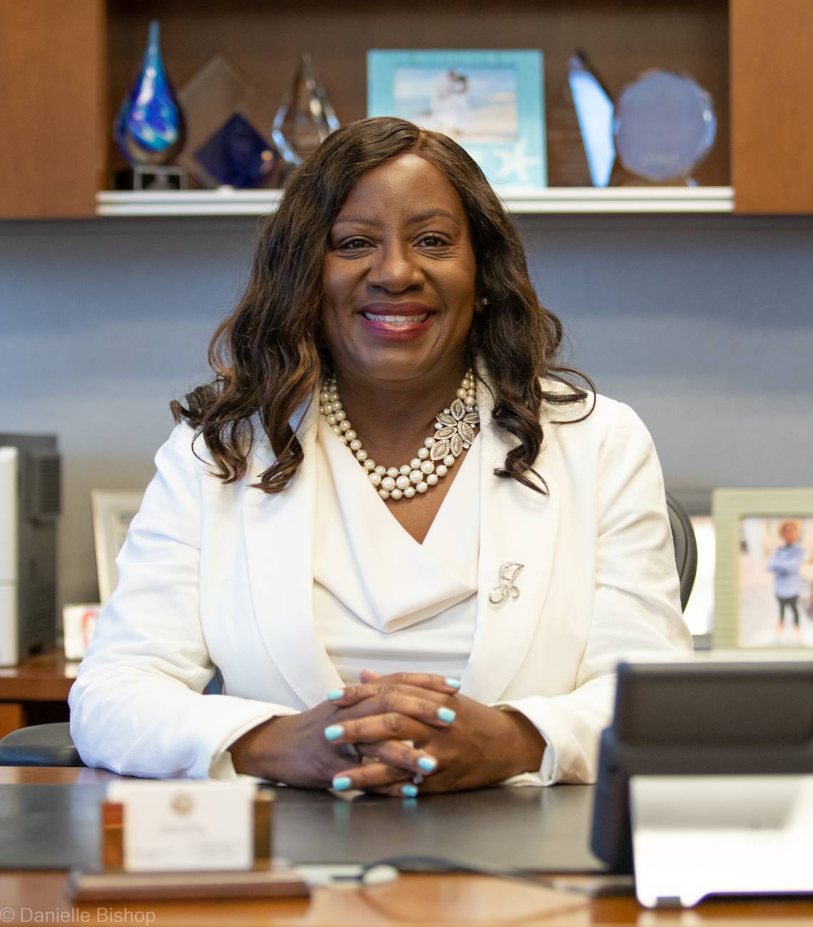 Judge Jackie Powell smiling at her desk in a white blazer and pearls, awards on the shelf behind her