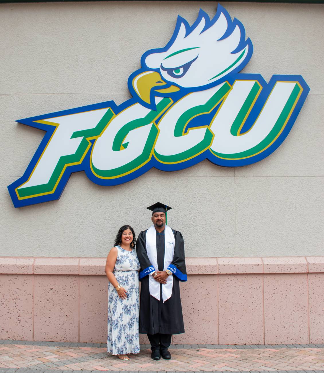 A graduate in cap and gown beside his mother at the FGCU eagle wall