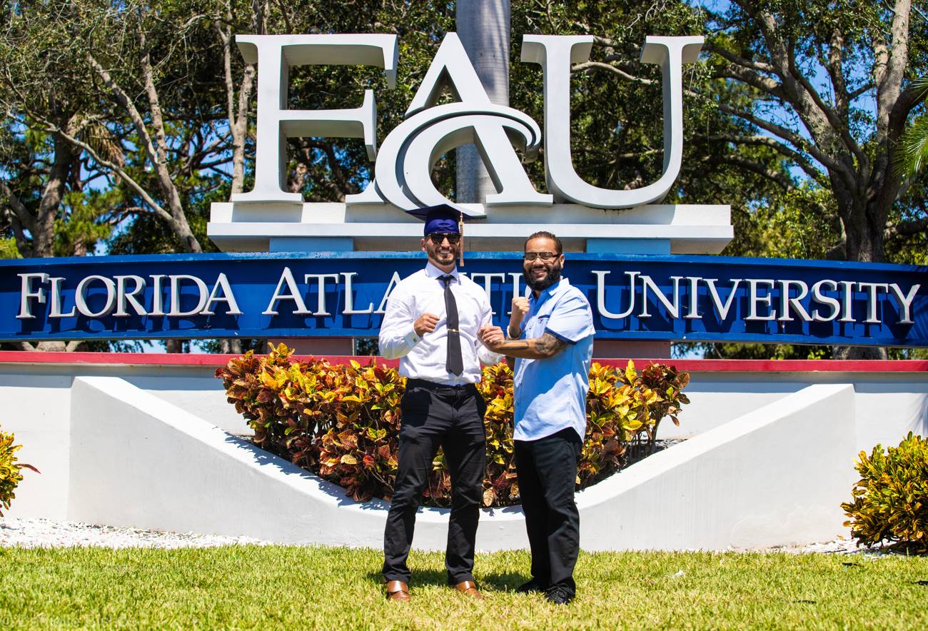 Two men flexing proudly in front of the Florida Atlantic University entrance sign