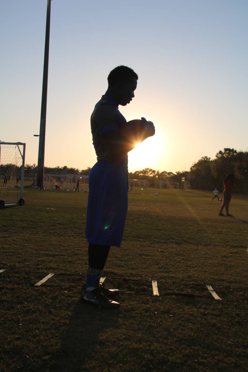 A young football player silhouetted against the setting sun, holding the ball close on the practice field