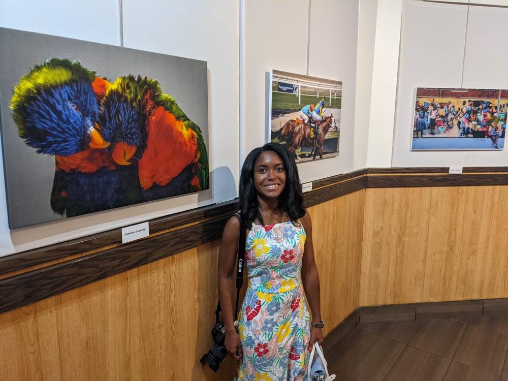 Danielle standing beside her exhibited prints at Island SPACE, including Compassion, the lorikeets canvas with her name card beneath it