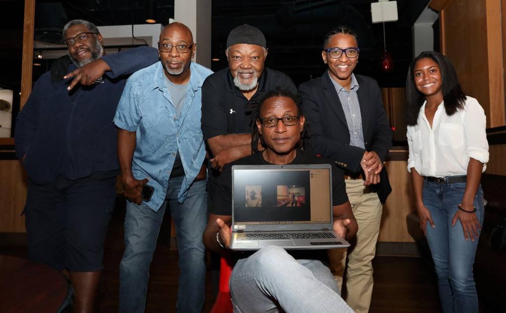 The Through the Caribbean Lens photographers and mentors gathered together, one holding a laptop with remote participants joining in