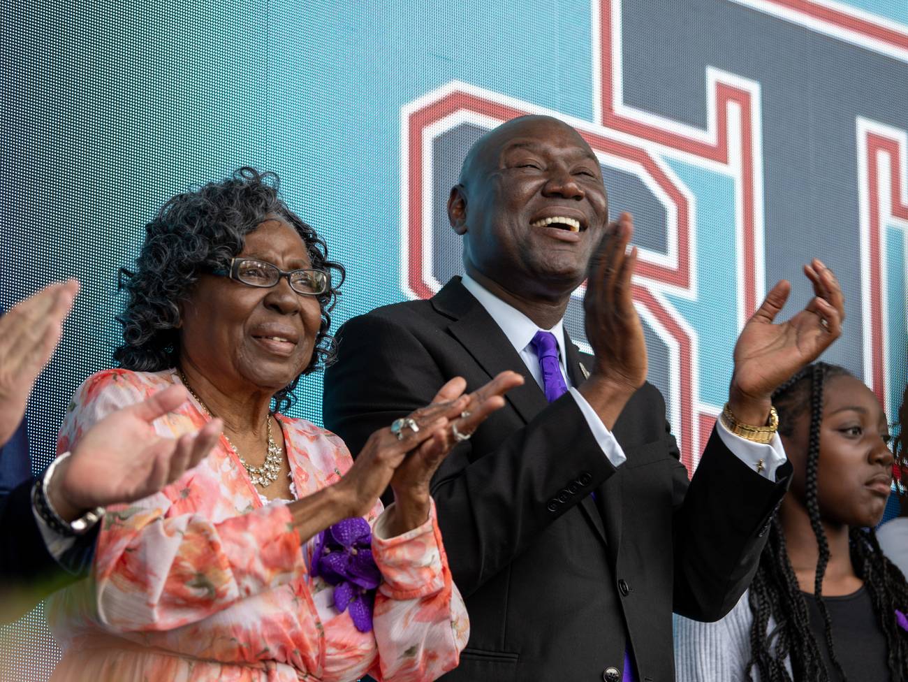 Ben Crump laughing and applauding on stage beside a woman in a floral dress at the dedication