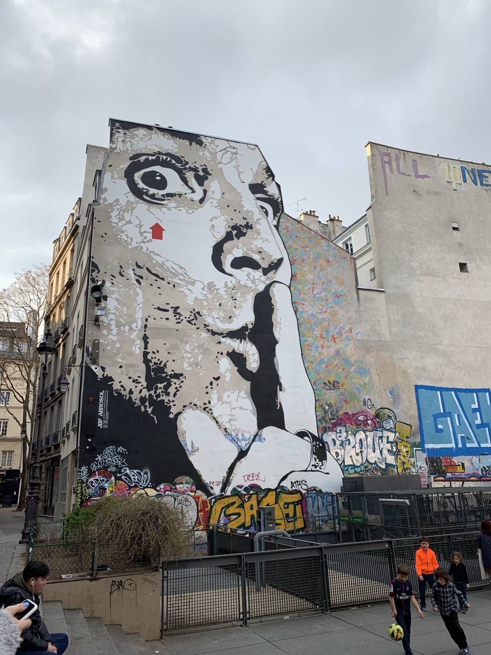 A towering black and white mural of a face shushing, surrounded by graffiti on a Paris street